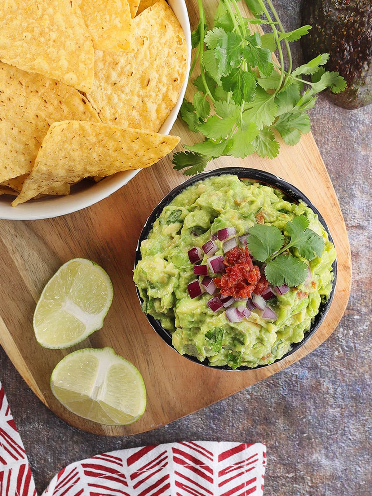 Chunky guacamole garnished with minced red onion, diced sun dried tomato, and cilantro with a bowl of tortilla chips, lime wedges, and cilantro on the side.