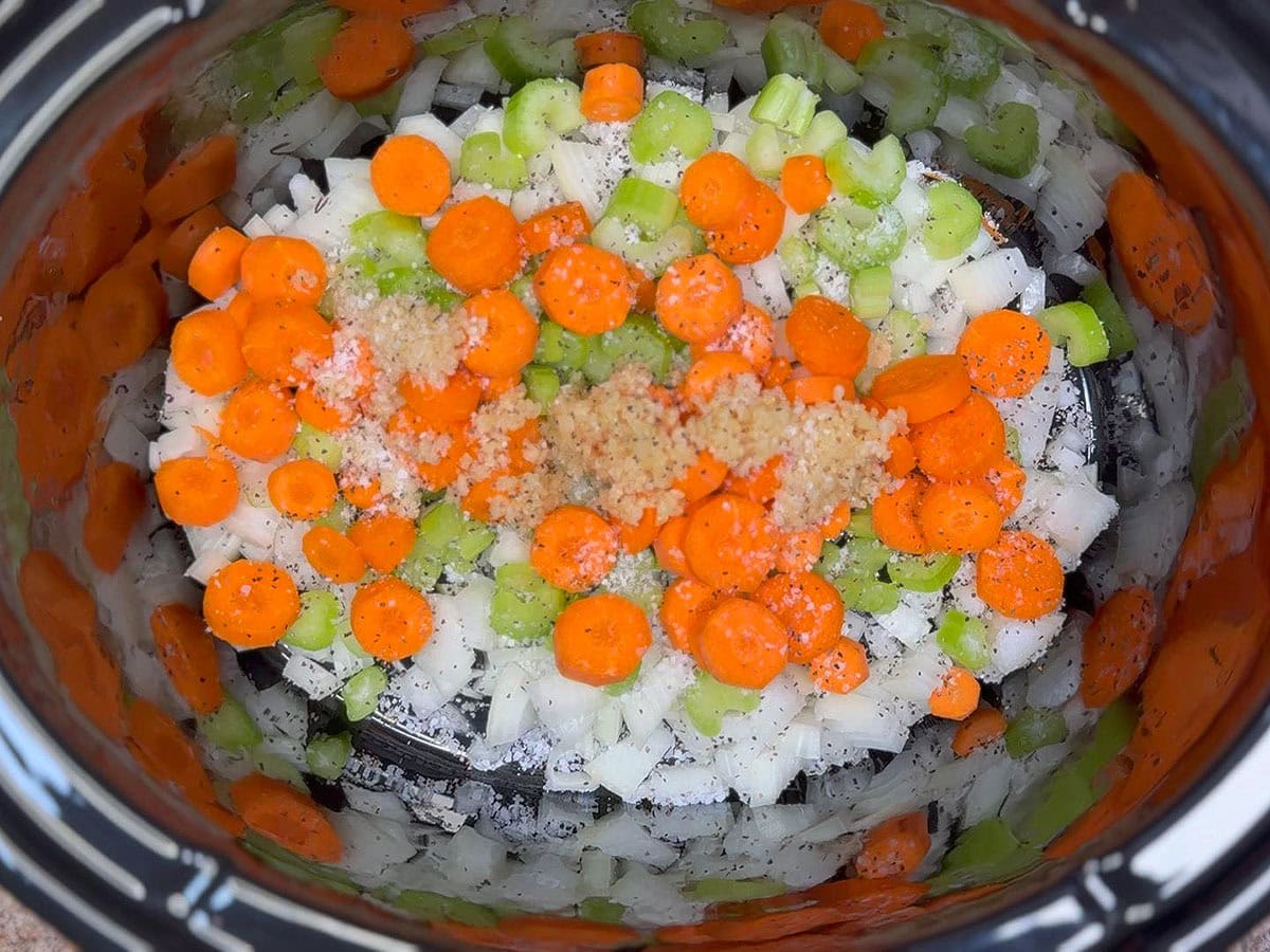 Vegetables and minced garlic seasoned with salt and pepper in the bottom of a slow cooker.