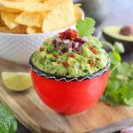 Chunky guacamole garnished with minced red onion, diced sun dried tomato, and cilantro with a bowl of tortilla chips in the background.