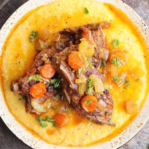 Slow cooker country style pork ribs nestles on a bed of cheese grits and topped with parsley, carrots, onions, and celery.