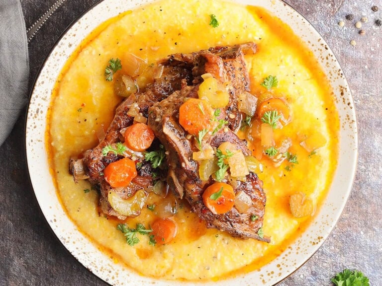 Slow cooker country style pork ribs nestles on a bed of cheese grits and topped with parsley, carrots, onions, and celery.