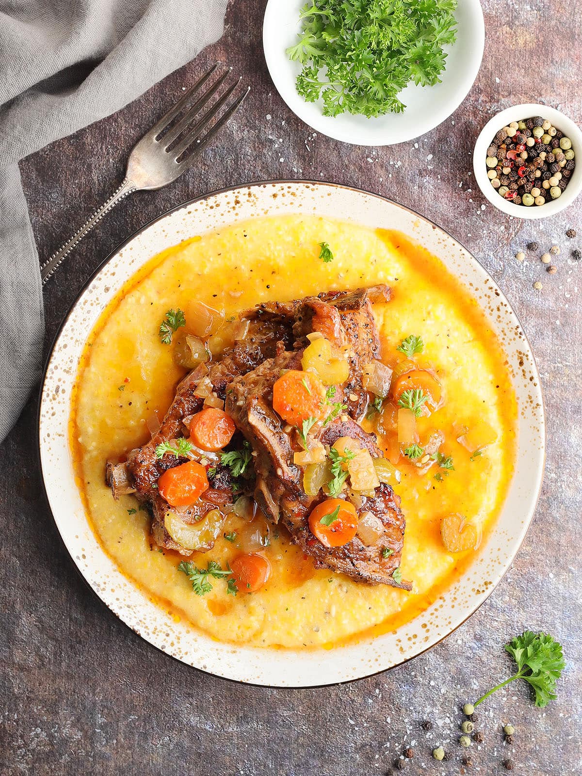 Slow cooker country style pork ribs nestles on a bed of cheese grits and topped with parsley, carrots, onions, and celery.