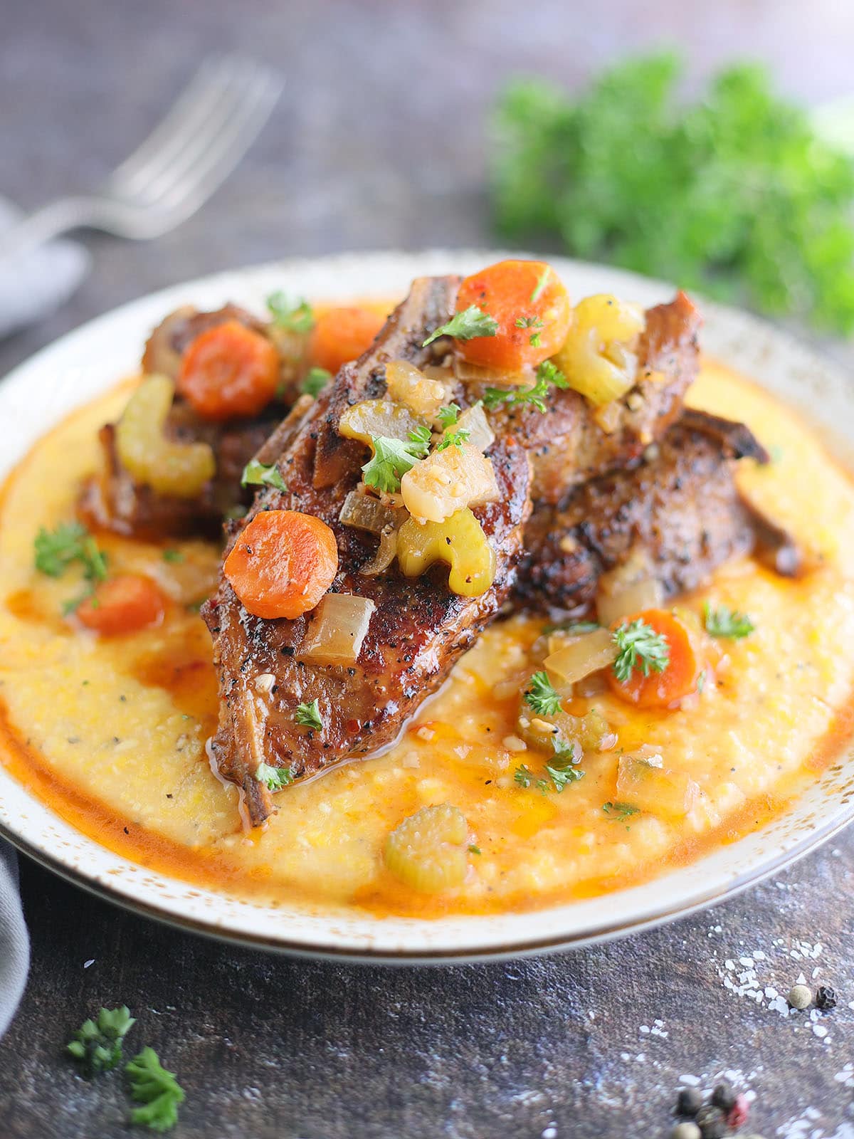 Slow cooker country style pork ribs nestles on a bed of cheese grits and topped with parsley, carrots, onions, and celery.