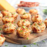 Bacon and cheese muffins garnished with fresh chives on a wooden cutting board with a wedge of cheese and cooked bacon in the background.