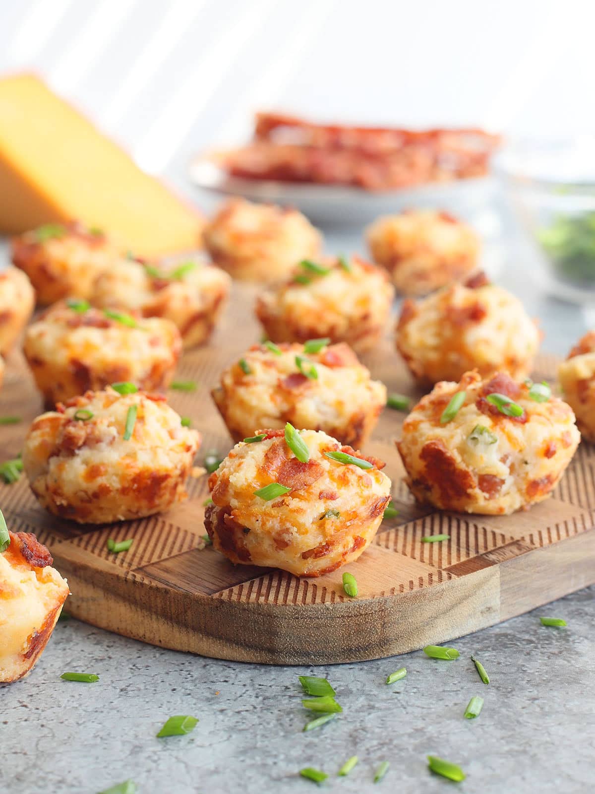 Bacon and cheese muffins garnished with fresh chives on a wooden cutting board with a wedge of cheese and cooked bacon in the background.