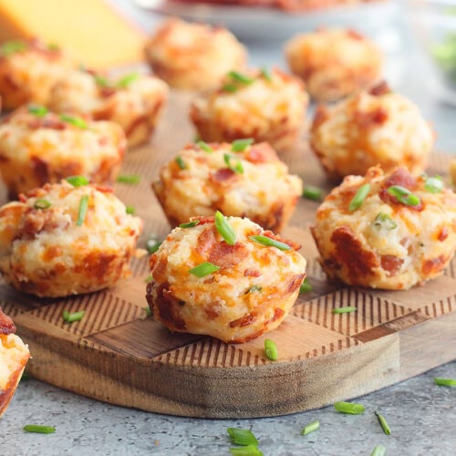 Bacon and cheese muffins garnished with fresh chives on a wooden cutting board with a wedge of cheese and cooked bacon in the background.
