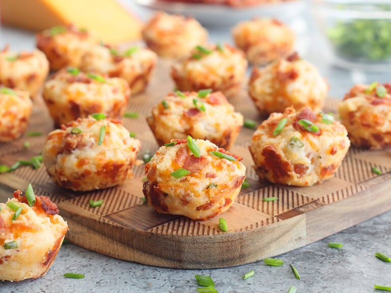 Bacon and cheese muffins garnished with fresh chives on a wooden cutting board with a wedge of cheese and cooked bacon in the background.