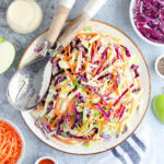 Serving dish of apple cabbage slaw with purple cabbage, shredded carrots, extra dressing and other ingredients arranged on the side.