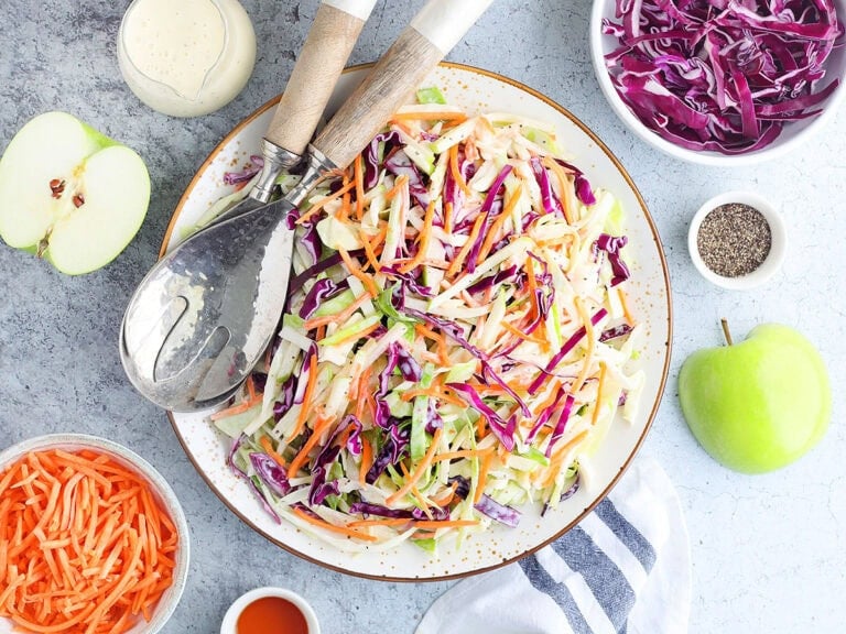 Serving dish of apple cabbage slaw with purple cabbage, shredded carrots, extra dressing and other ingredients arranged on the side.