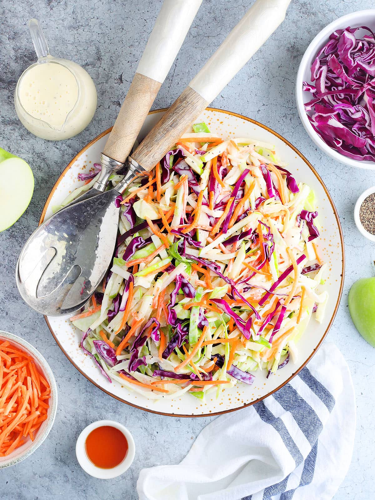 Serving dish of apple cabbage slaw with purple cabbage, shredded carrots, extra dressing and other ingredients arranged on the side.