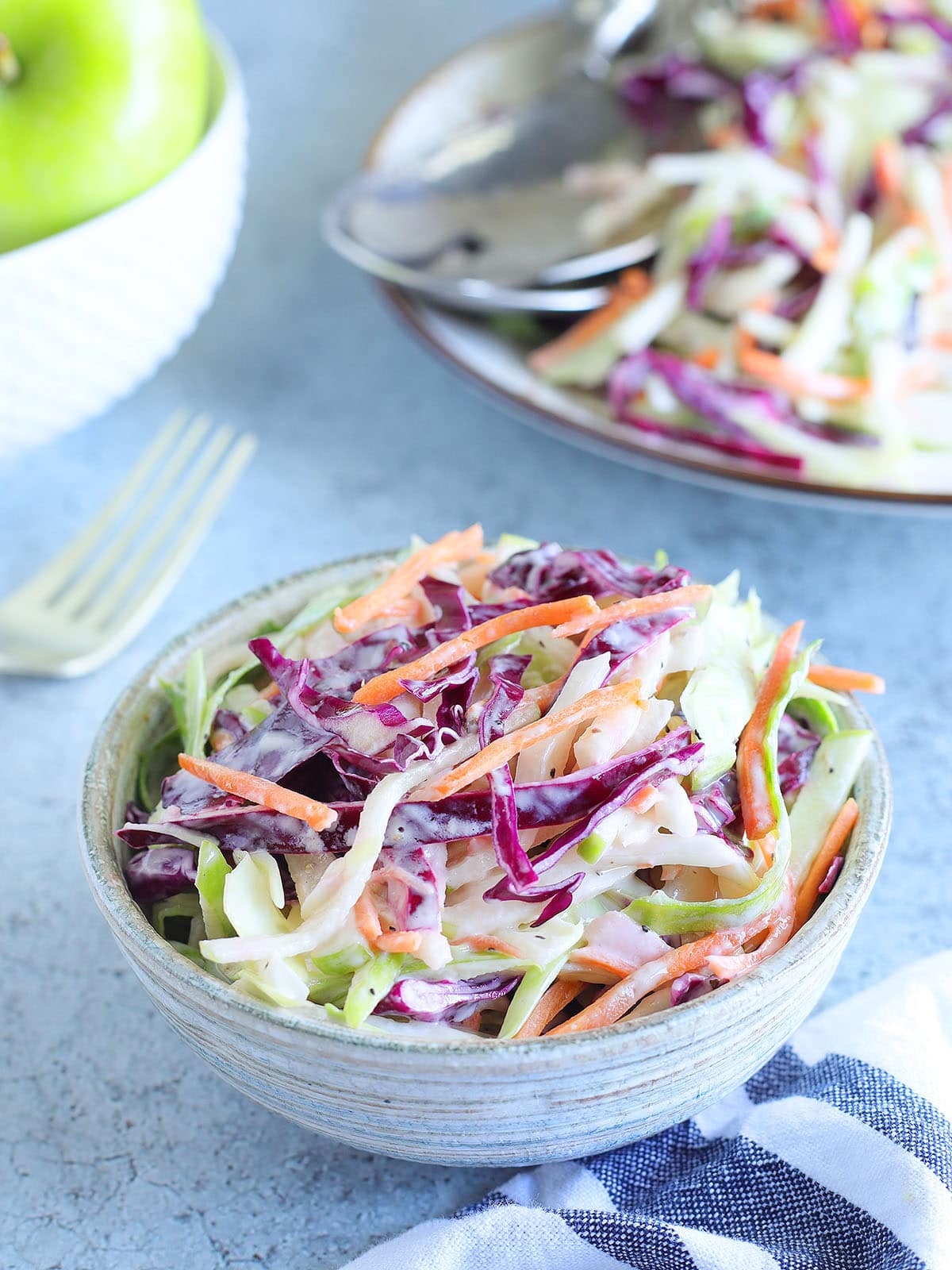 Dish of apple and cabbage slaw with a bowl of green apples and a serving bowl of slaw in the background.