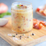 Jar of hot bacon dressing on a wooden cutting board with a sliced shallot and bacon slices in the background.