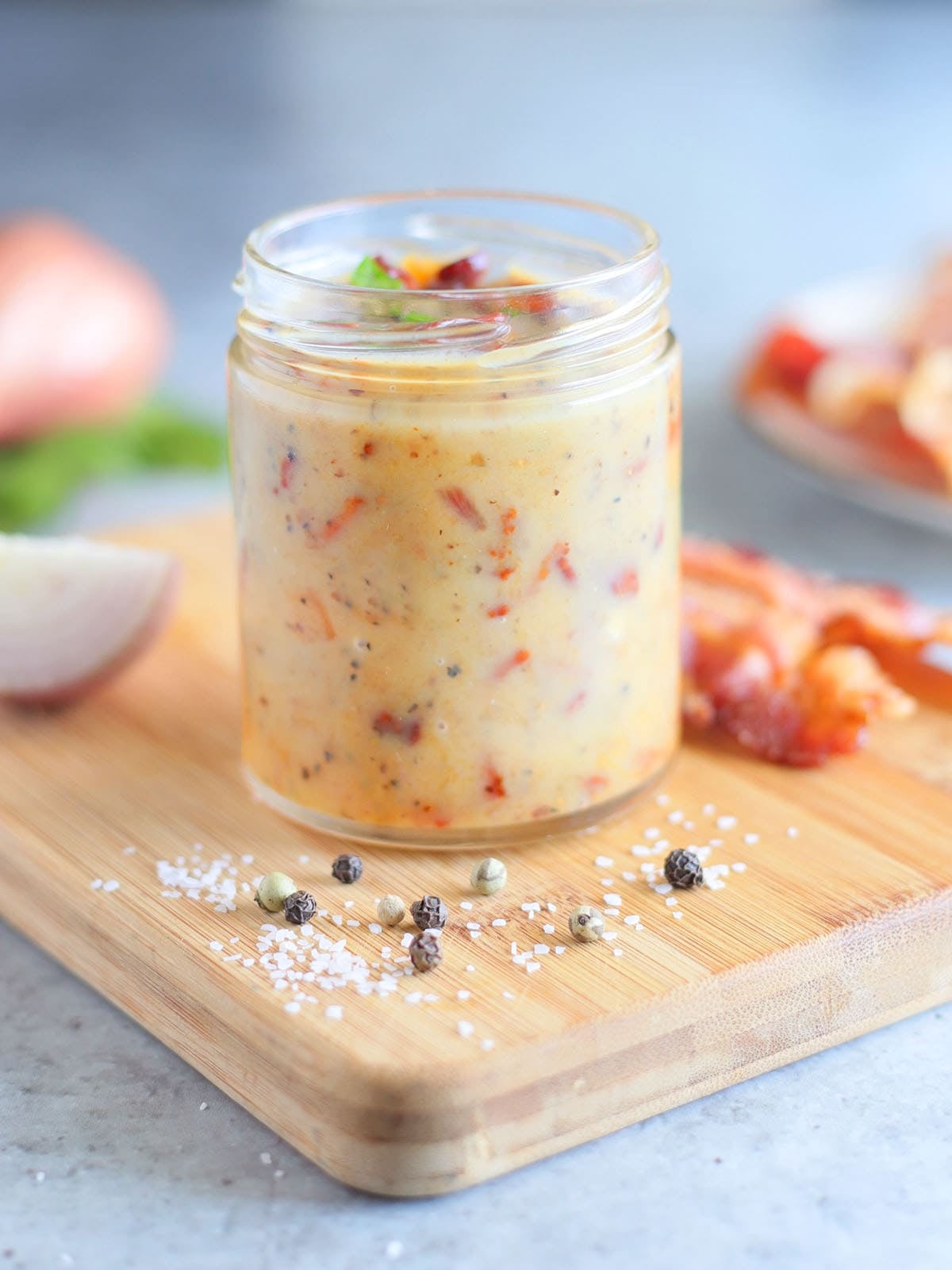 Jar of hot bacon dressing on a wooden cutting board with a sliced shallot and bacon slices in the background.