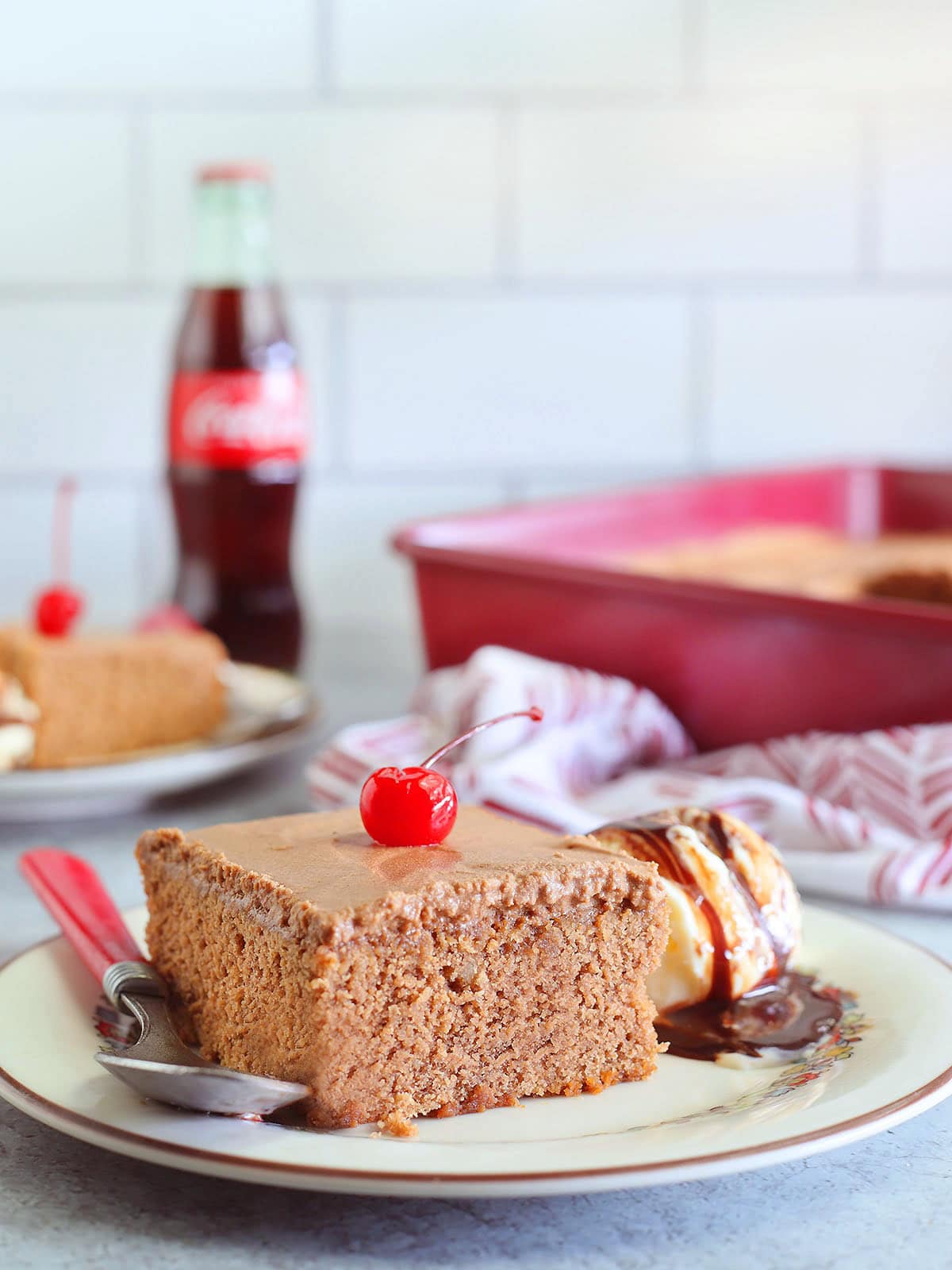 Slice of chocolate coca cola cake garnished with a cherry and a scoop of vanilla ice cream with chocolate syrup.