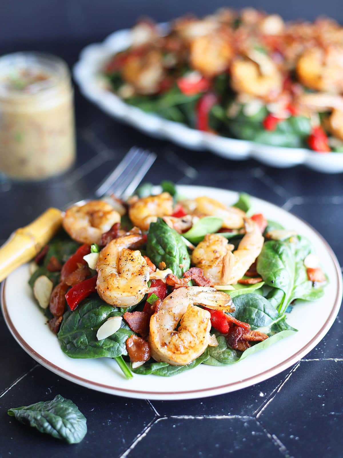 Plated grilled shrimp salad with a fork on the side, a jar of hot bacon dressing and platter of full salad in the background.