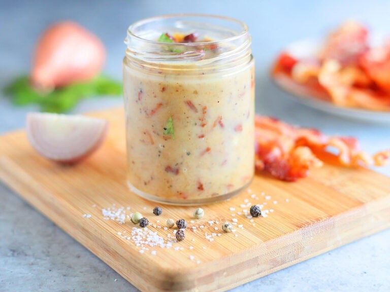 Jar of hot bacon dressing on a wooden cutting board with a sliced shallot and bacon slices in the background.