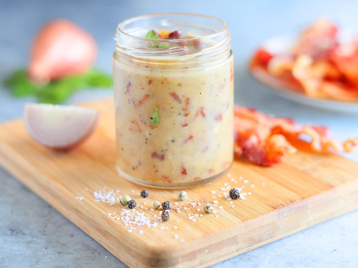 Jar of hot bacon dressing on a wooden cutting board with a sliced shallot and bacon slices in the background.