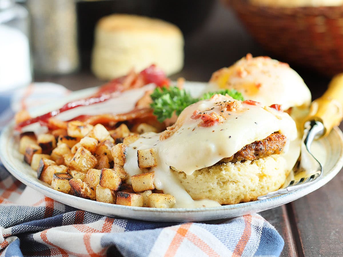 Country eggs benedict on a plate with a side of hash brown potatoes and bacon.