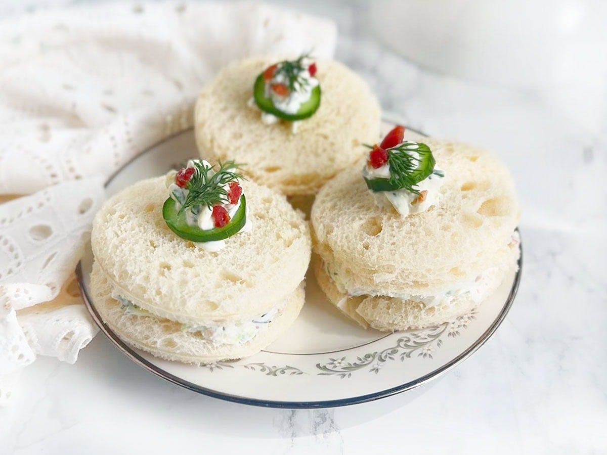 Three cucumber tea sandwiches garnished with a dollop of cream cheese, cucumber slice, fresh dill and bacon bits.