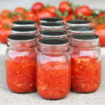 Several glass mason jars filled with diced tomatoes with a pile of fresh whole tomatoes in the background.