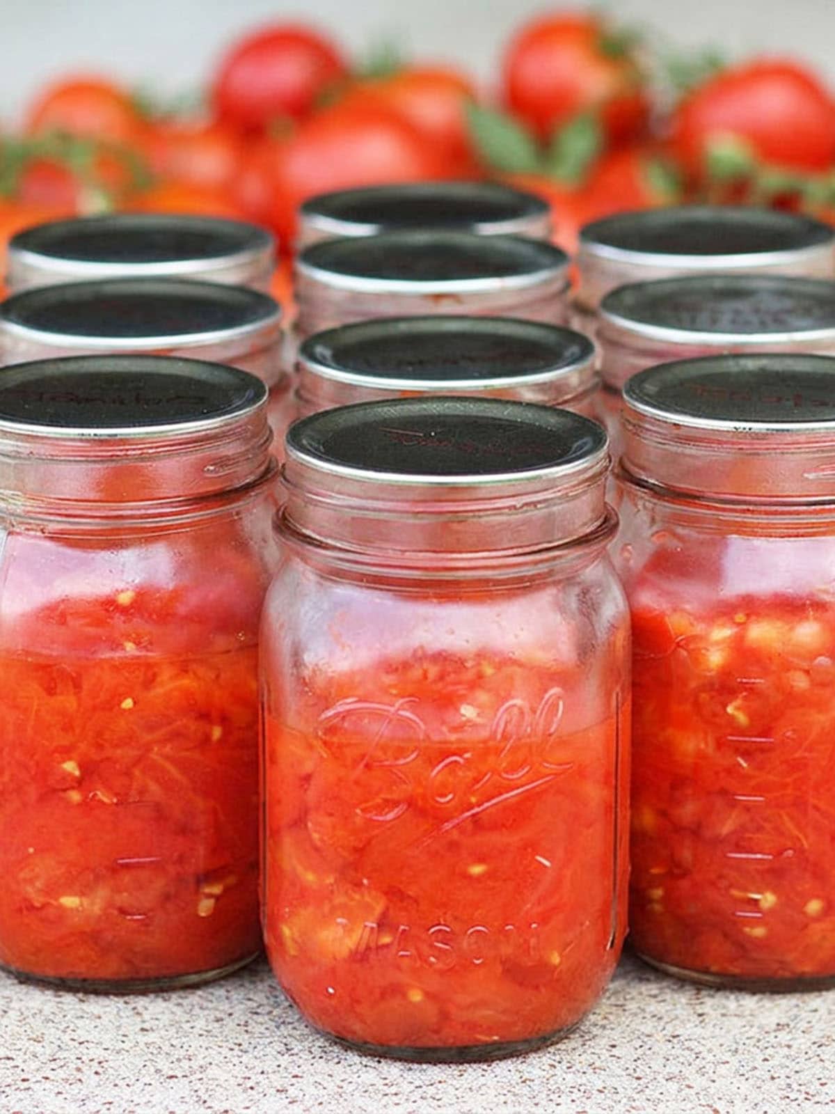 Several glass mason jars filled with diced tomatoes with a pile of fresh whole tomatoes in the background.