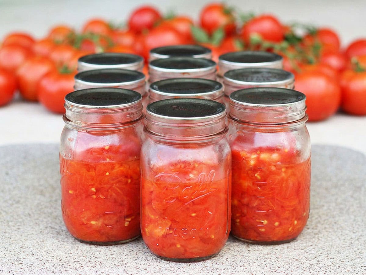 Several glass mason jars filled with diced tomatoes with a pile of fresh whole tomatoes in the background.