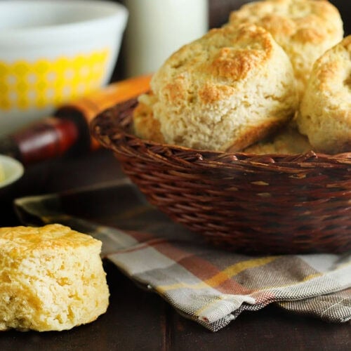 A basket of biscuits with steam rising from the tops, a single biscuit in the corner, with a mixing bowl, a small dish of butter and a bottle of milk in the background.
