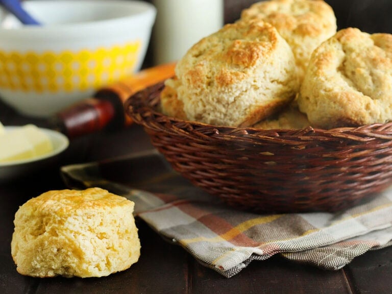 A basket of biscuits with steam rising from the tops, a single biscuit in the corner, with a mixing bowl, a small dish of butter and a bottle of milk in the background.