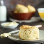 A single buttermilk biscuit on a plate with a knife topped with a pat of warm butter. A basket of biscuits, butter, and a bottle of milk are in the background.