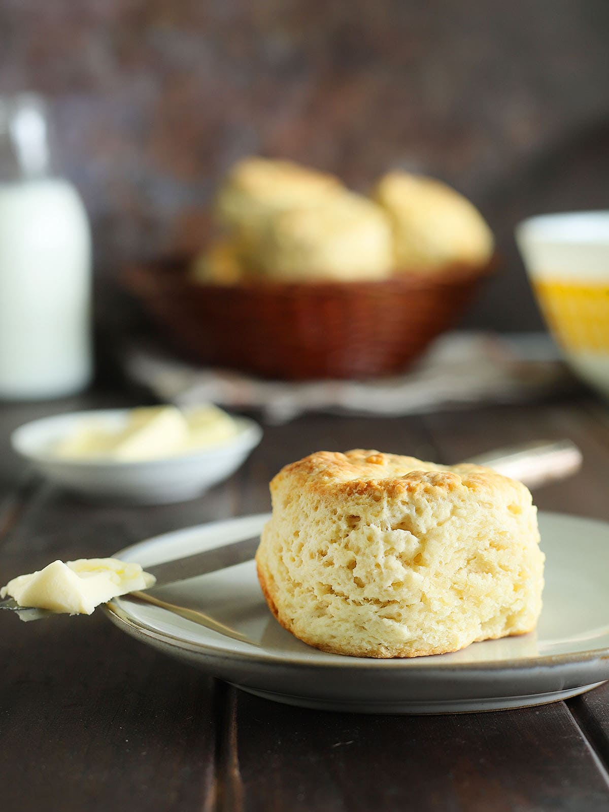 A single buttermilk biscuit on a plate with a knife topped with a pat of warm butter. A basket of biscuits, butter, and a bottle of milk are in the background.