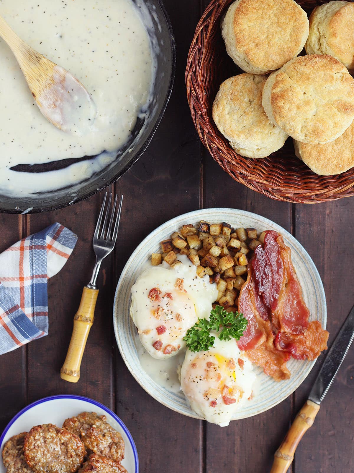 Country eggs benedict on a plate with a side of hash brown potatoes and bacon. A pan of cream gravy, a basket of biscuits, and a small serving plate of patty sausage can be seen on the side.