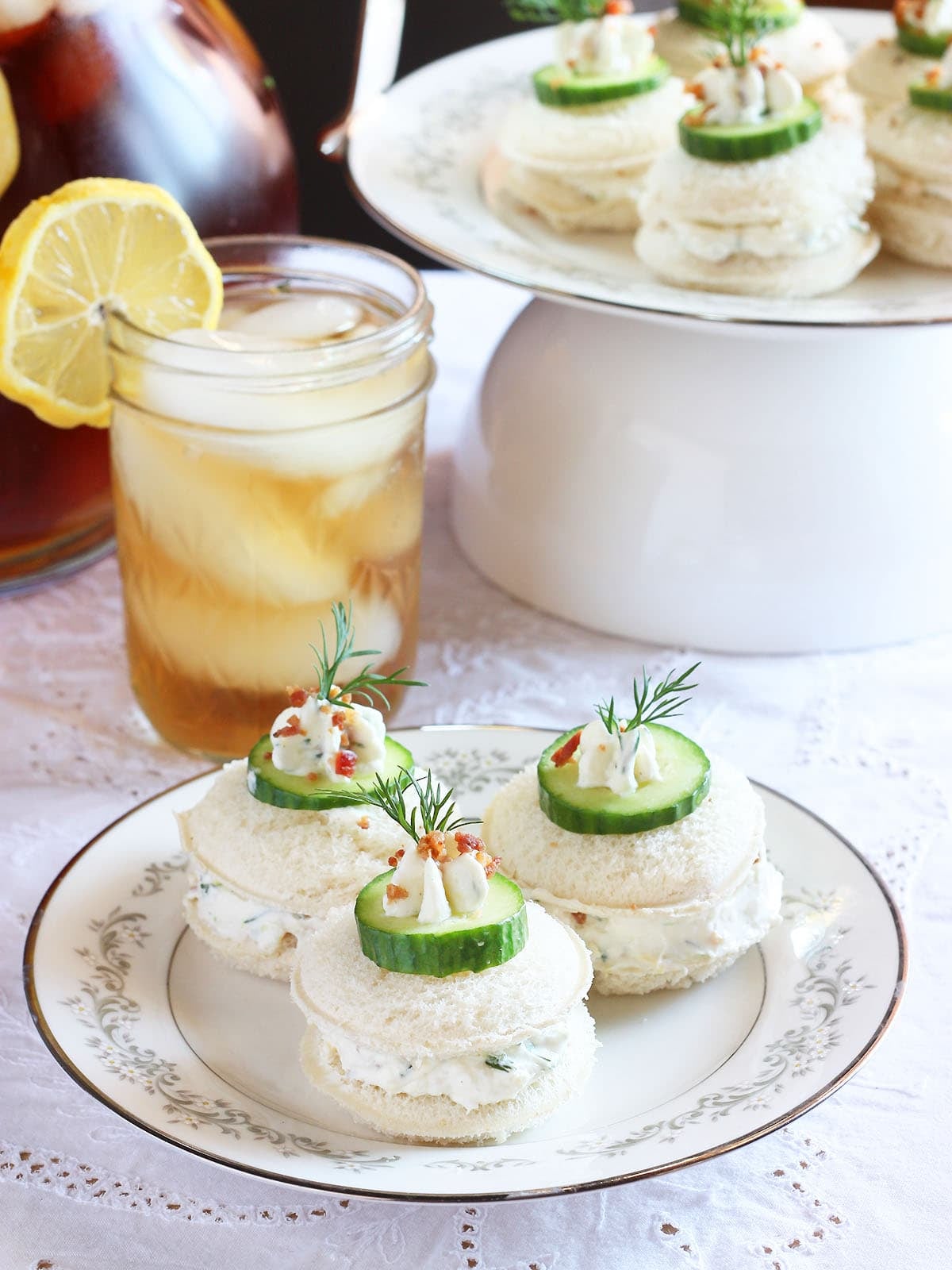 Three cucumber tea sandwiches garnished with a dollop of cream cheese, cucumber slice, fresh dill and bacon bits. Platter of sandwiches and a glass of iced tea in the background.