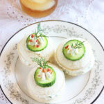 Three cucumber tea sandwiches garnished with a dollop of cream cheese, cucumber slice, fresh dill and bacon bits. Glass of iced tea in the background.