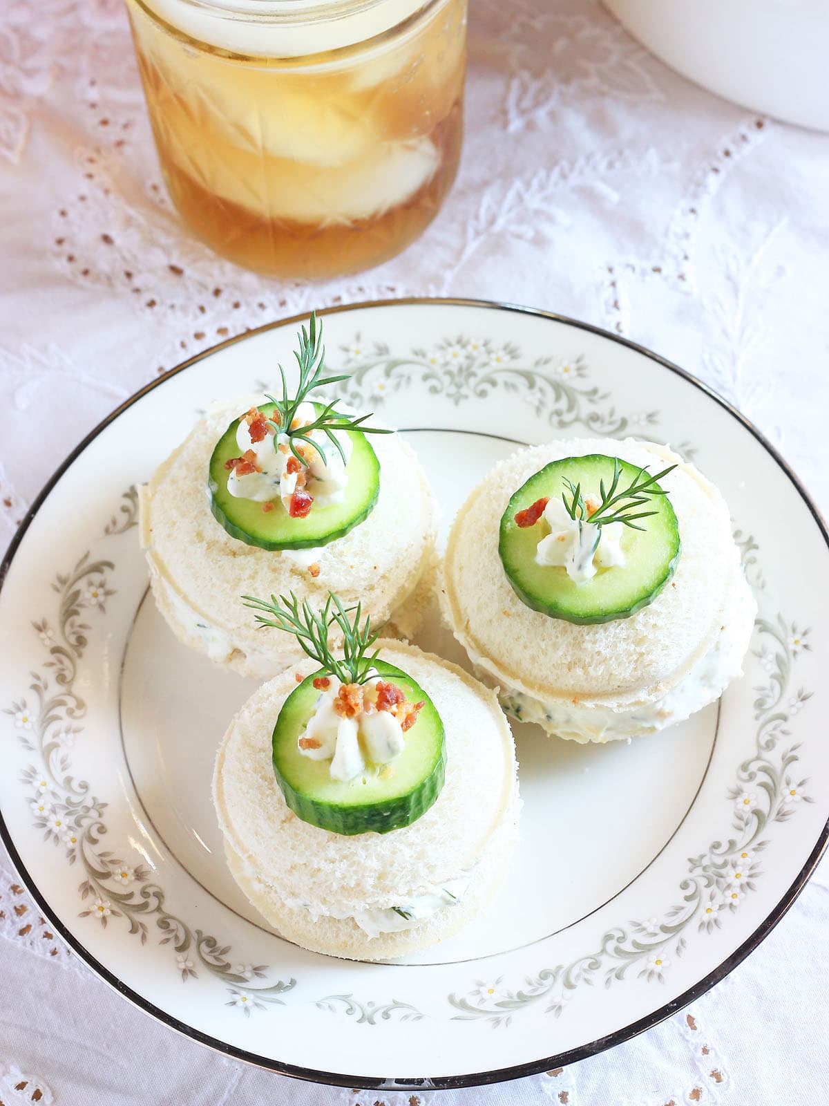 Three cucumber tea sandwiches garnished with a dollop of cream cheese, cucumber slice, fresh dill and bacon bits. Glass of iced tea in the background.