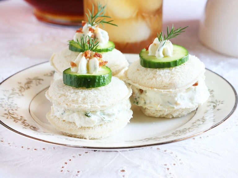 Three cucumber tea sandwiches garnished with a dollop of cream cheese, cucumber slice, fresh dill and bacon bits.