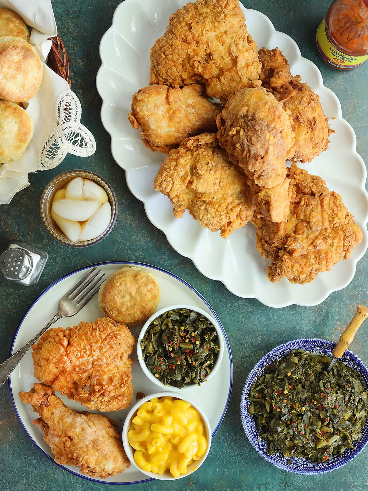 Table set with a platter of fried chicken, a basket of biscuits, a serving bowl of collard greens, and a glass of iced tea. A plate with a serving of chicken, macaroni, and collard greens to the side.