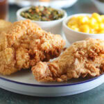 Two pieces of fried chicken on a plate with a dish of macaroni and cheese and collard greens.