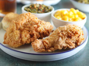 Two pieces of fried chicken on a plate with a dish of macaroni and cheese and collard greens.