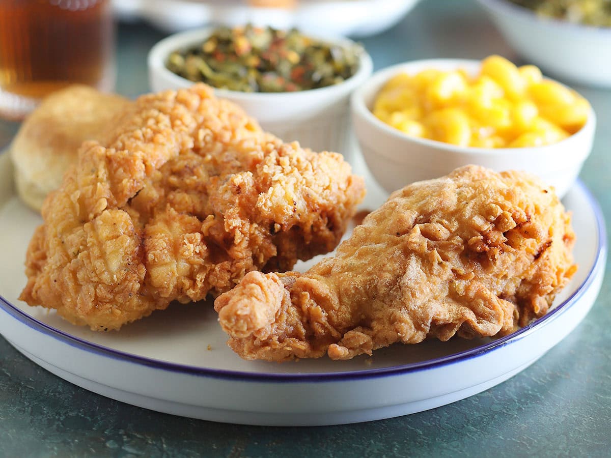 Two pieces of fried chicken on a plate with a dish of macaroni and cheese and collard greens.