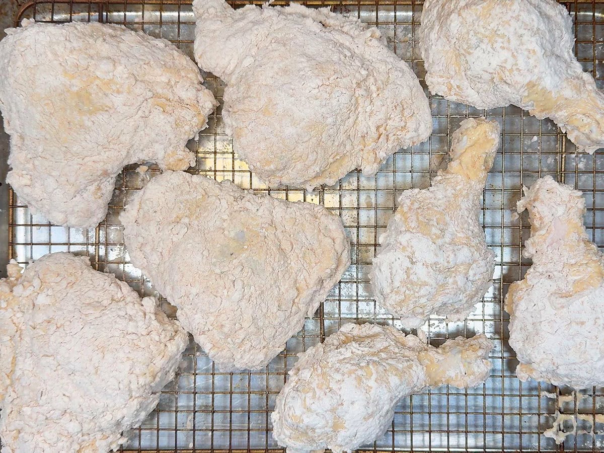 Battered and floured chicken thighs and legs resting on a wire rack before frying.