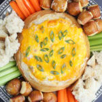 Baked Mississippi Sin Dip garnished with sliced green onions and served with torn bread, pretzel bites, carrot sticks, and celery sticks.