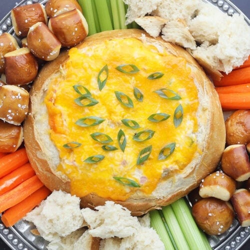 Baked Mississippi Sin Dip garnished with sliced green onions and served with torn bread, pretzel bites, carrot sticks, and celery sticks.