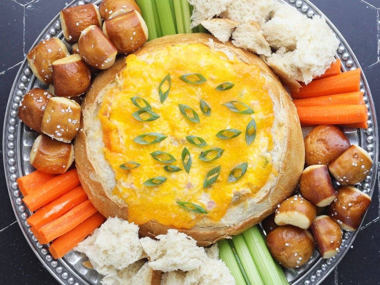 Baked Mississippi Sin Dip garnished with sliced green onions and served with torn bread, pretzel bites, carrot sticks, and celery sticks.
