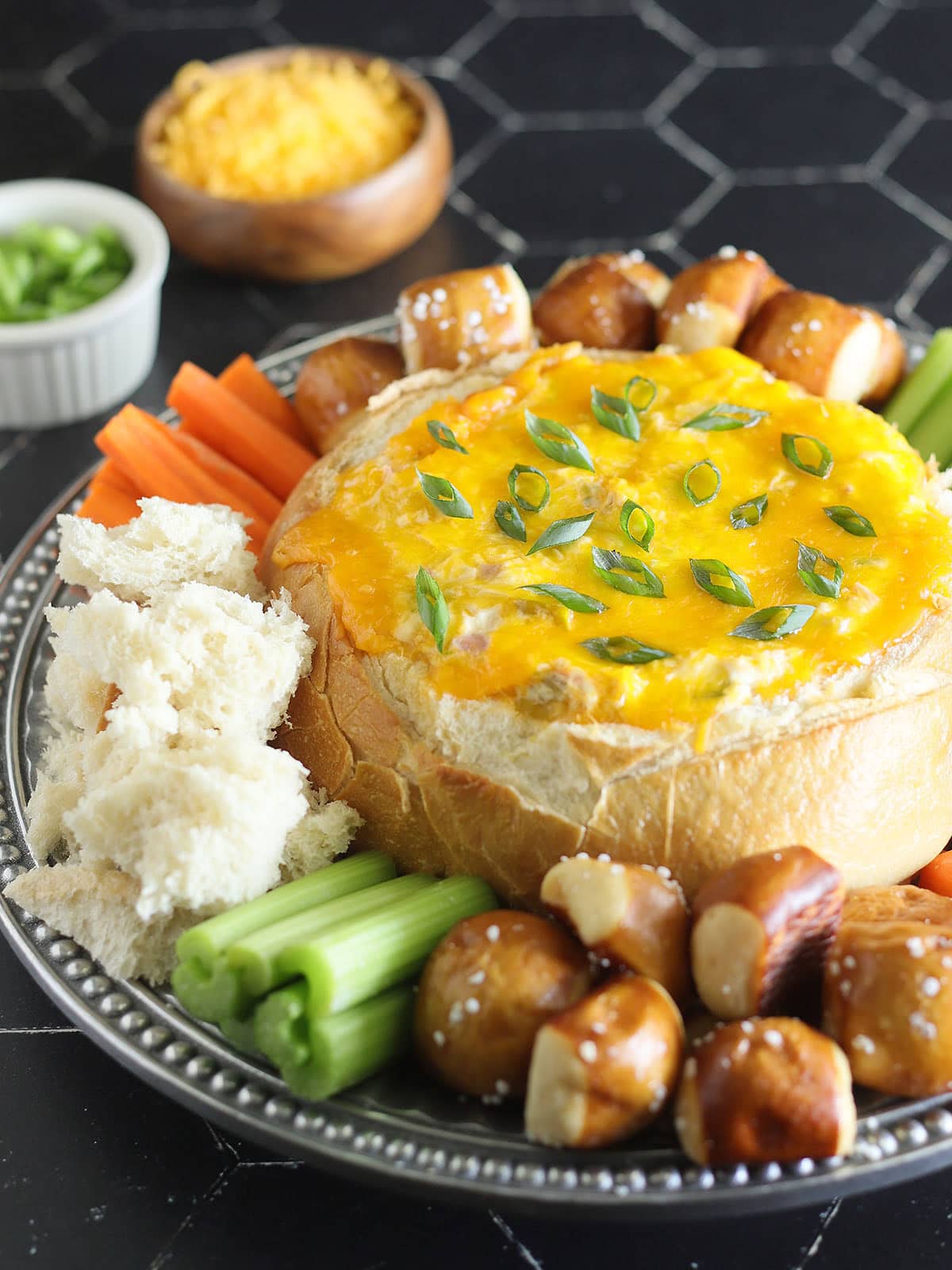 Baked Mississippi Sin Dip garnished with sliced green onions and served with torn bread, pretzel bites, carrot sticks, and celery sticks.