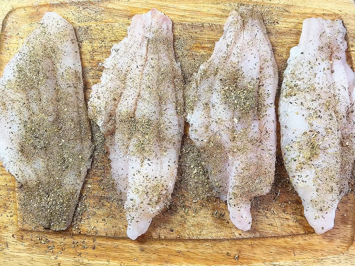 Four catfish fillets covered in seasoning laid out on a wooden cutting board.