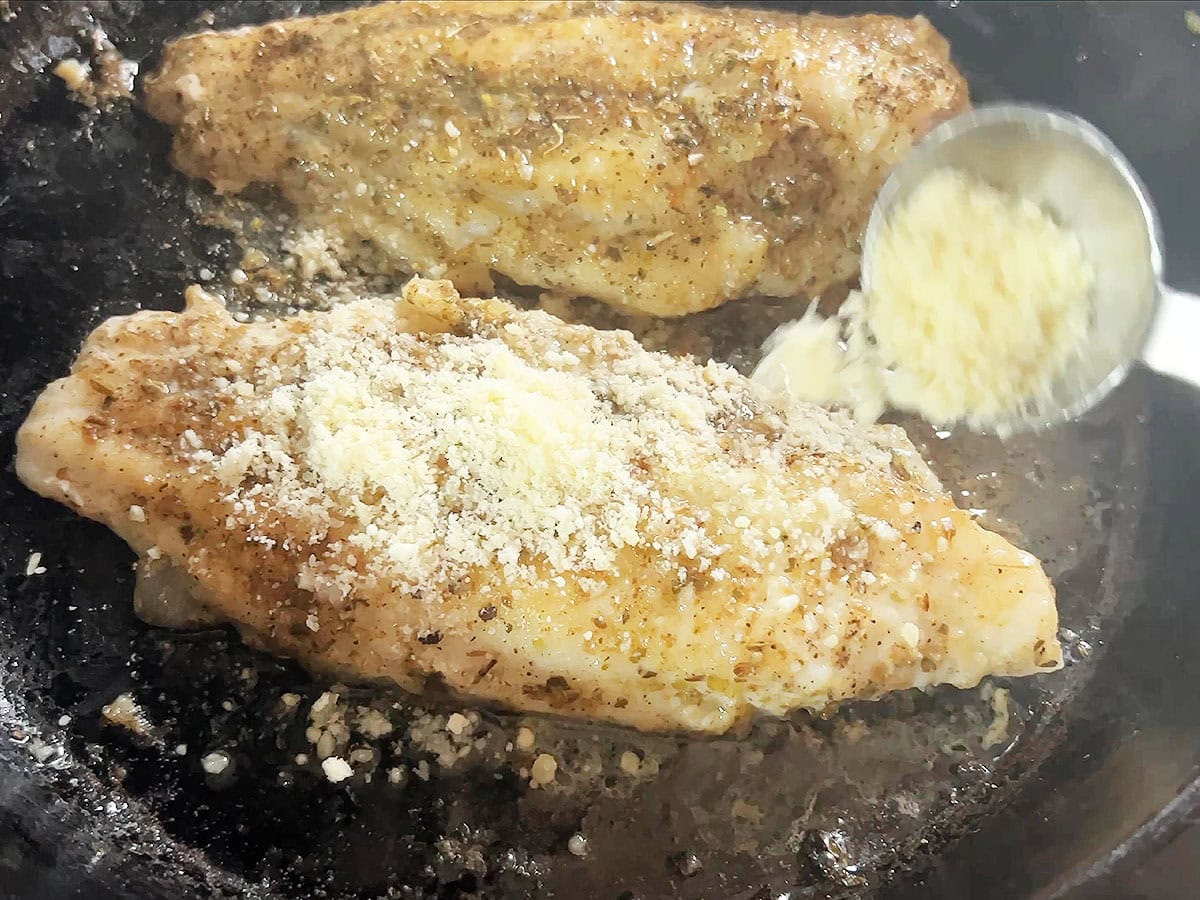 Sprinkling Parmesan cheese over two catfish fillets in a skillet.