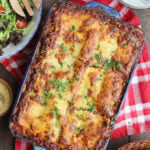 Pan of baked sausage lasagna with a bowl of fresh salad greens, garlic knots, and a dish of grated Parmesan cheese on the side.