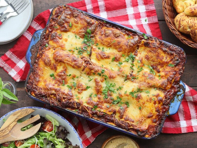 Pan of baked sausage lasagna with a bowl of fresh salad greens, garlic knots, and a dish of grated Parmesan cheese on the side.