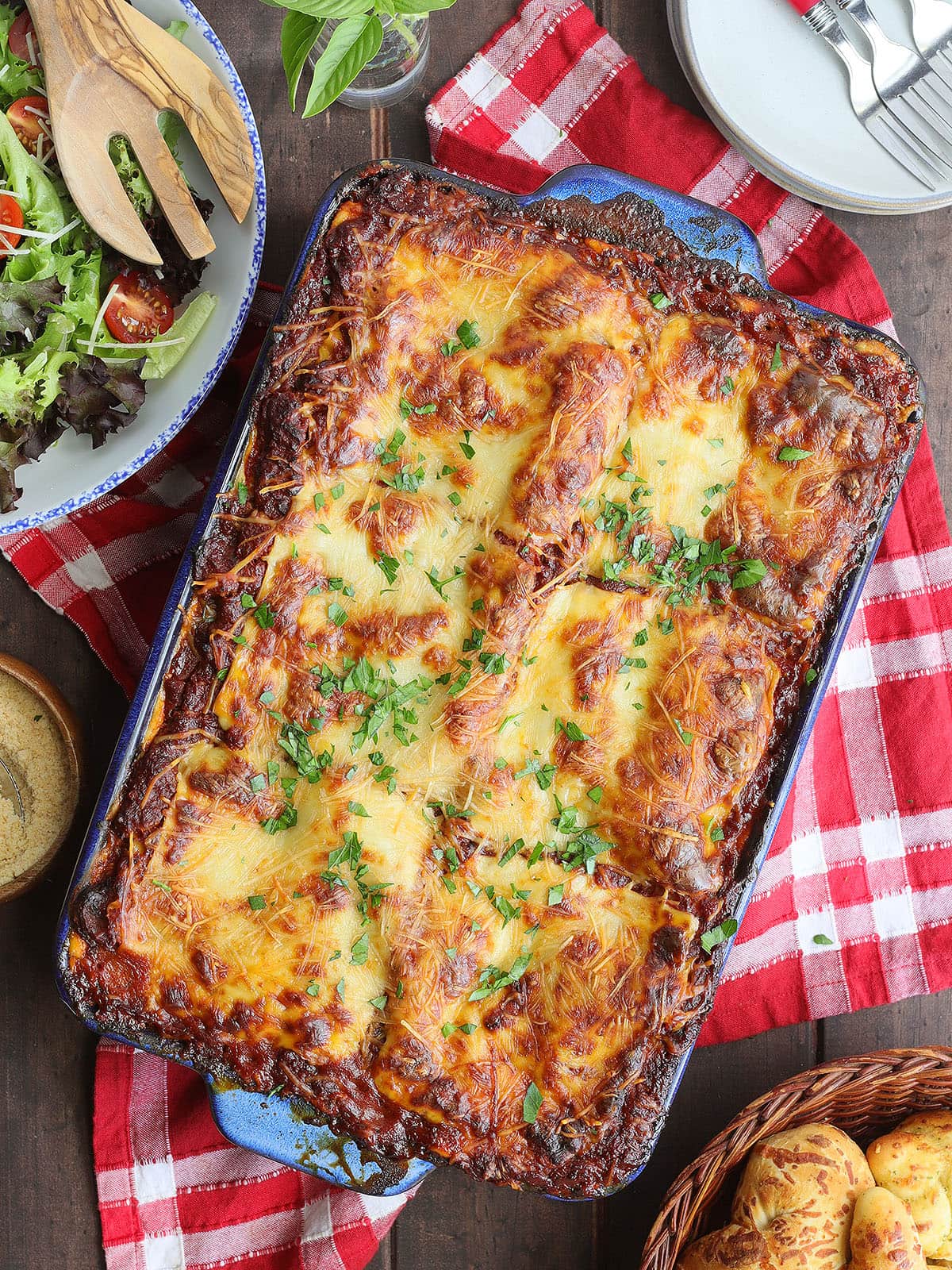 Pan of baked sausage lasagna with a bowl of fresh salad greens, garlic knots, and a dish of grated Parmesan cheese on the side.