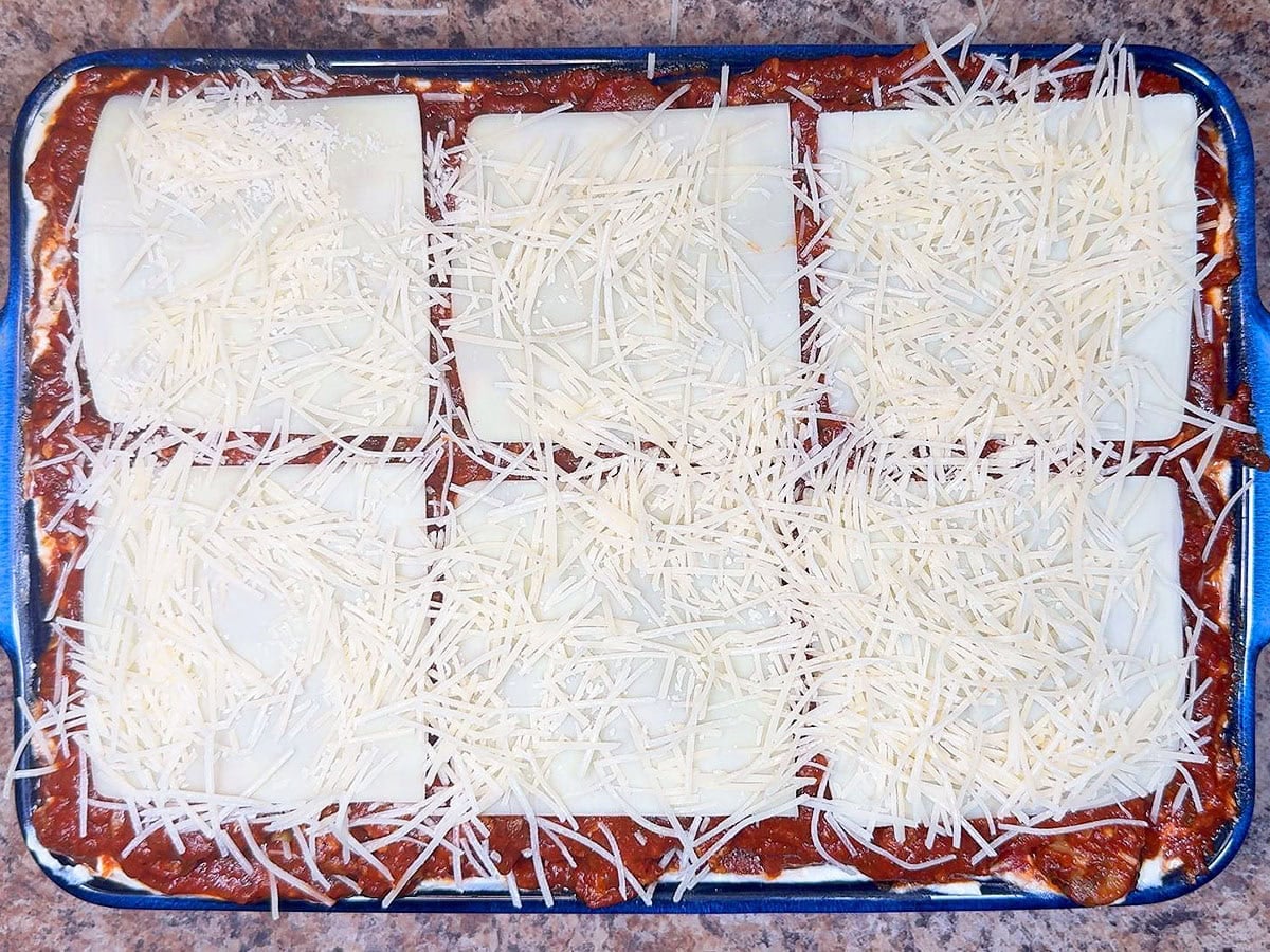 Unbaked sausage lasagna topped with meat sauce, mozzarella cheese slices, and shredded Parmesan cheese.